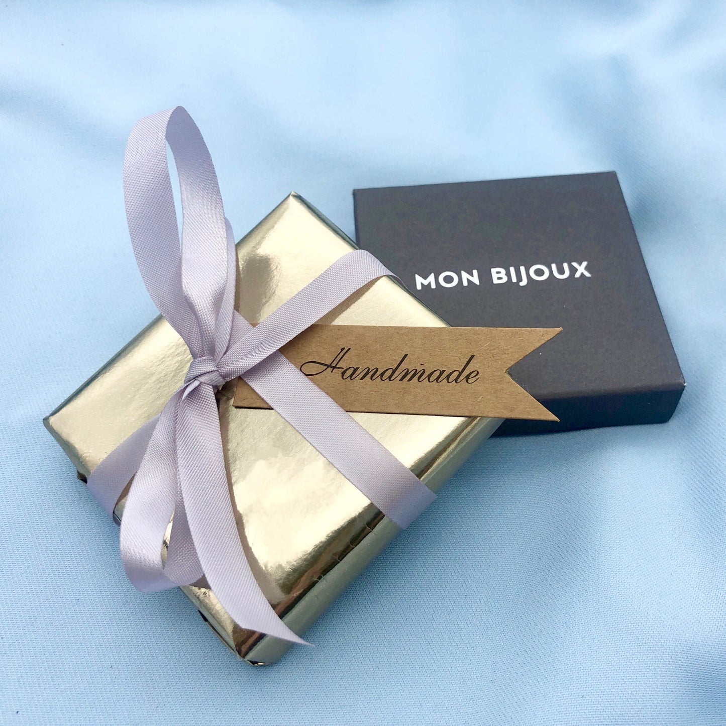 Gold jewelry box with a white ribbon and 'Mon Bijoux' packaging on a light blue background